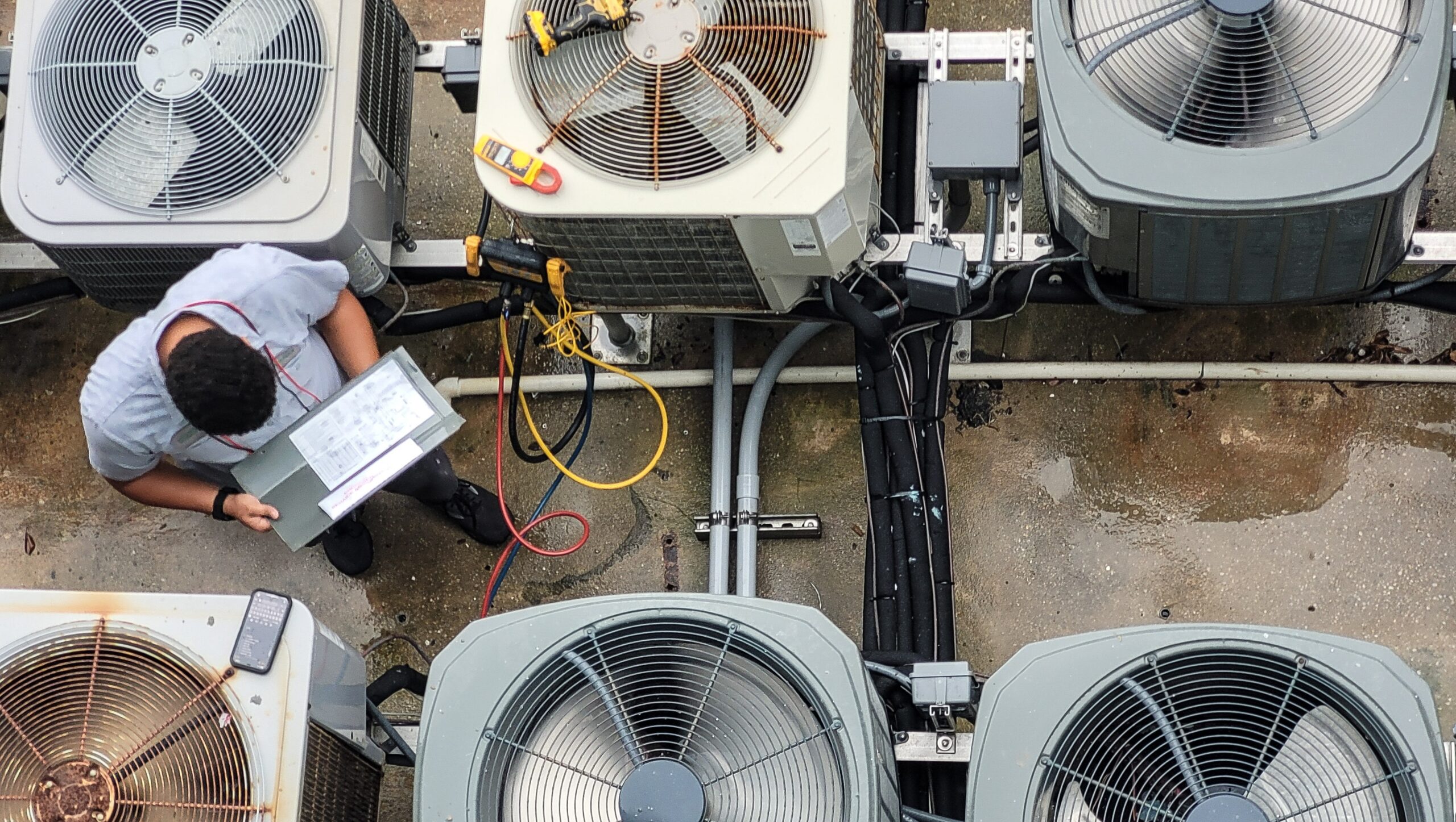 A technician performs HVAC System Inspection from above, checking air conditioning unit connections and readings for proper maintenance and repairs.